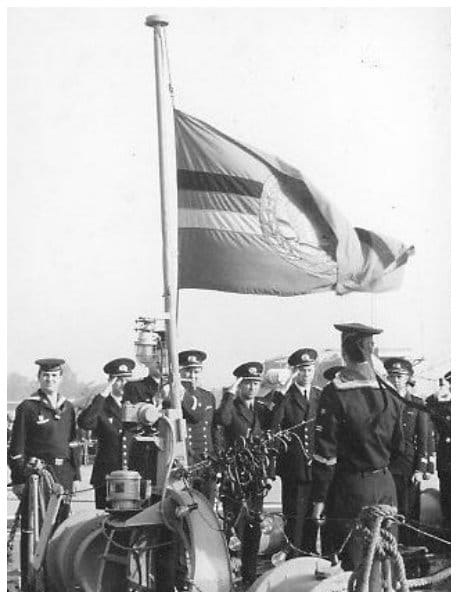 Flaggenparade an Deck: Offiziere und Matrosen stehen salutierend am Flaggenstock, an dem die Dienstflagge der Volksmarine im Wind steht