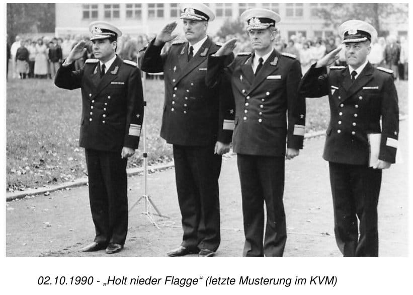 Vier Marineoffiziere stehen salutierend nebeneinander bei einer Musterung im Freien, im Hintergrund Zuschauer vor einem Gebäude